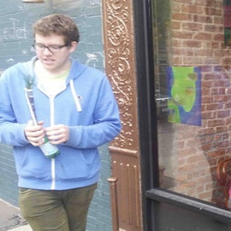 Jon Henry white man stands in front of window wearing blue sweatshirt and green pants and glasses