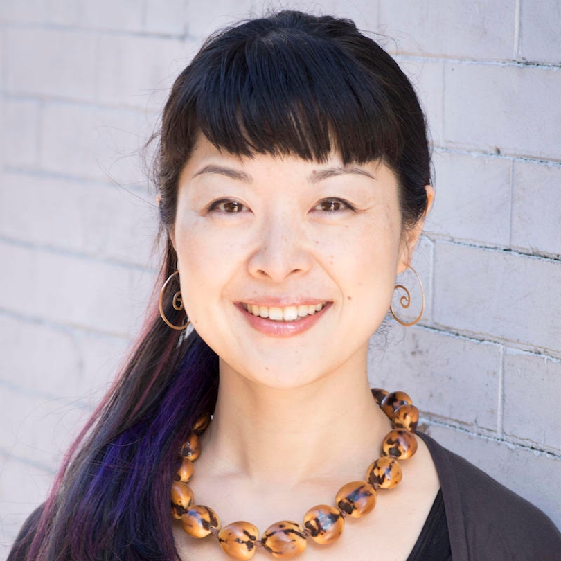 Miho Tsujii headshot Japanese woman with dark hair with bangs, orange necklace and dark shirt