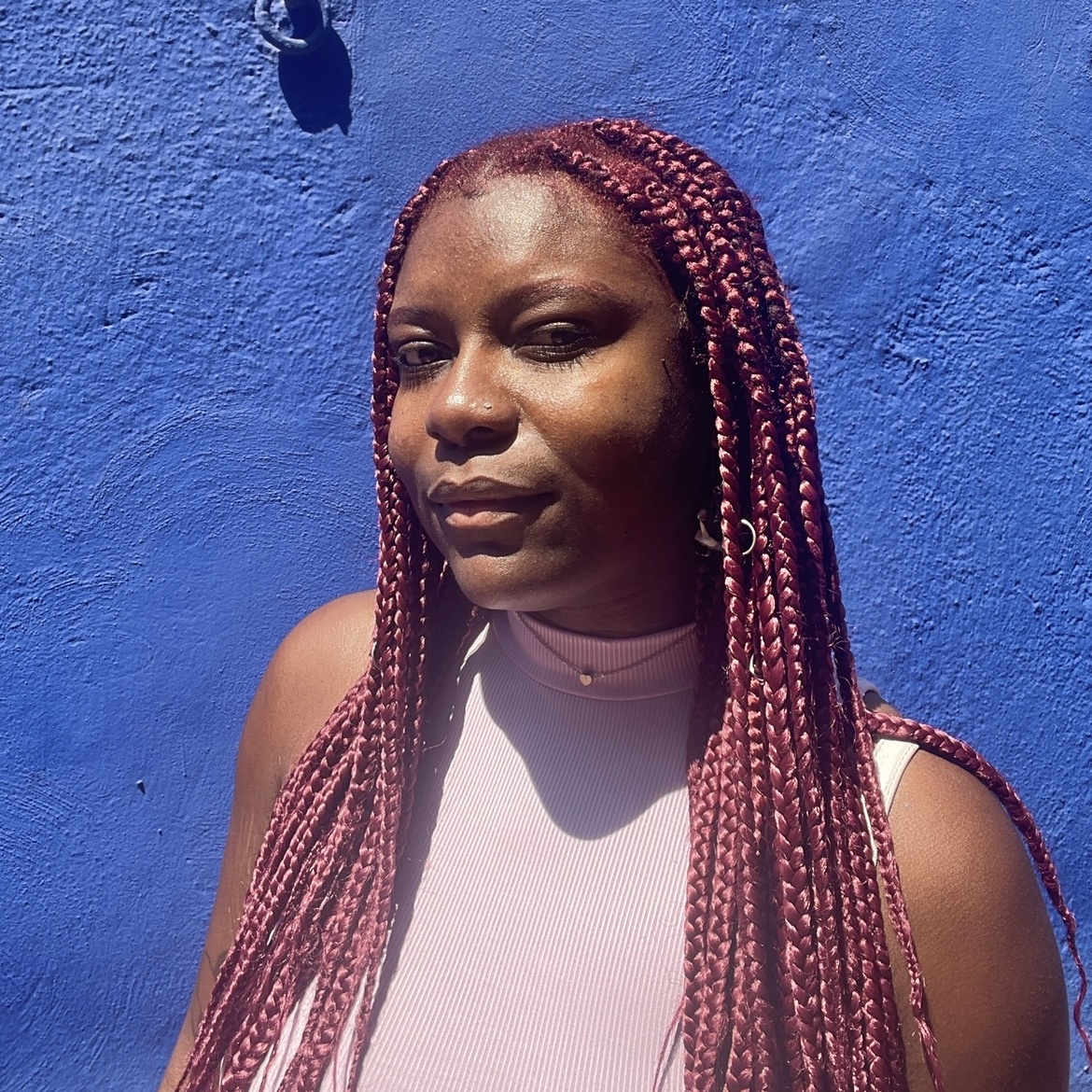 Edgie stands against a blue wall. She has long pink braids and a pink sleeveless turtleneck