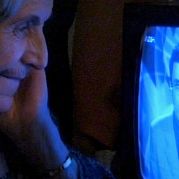 An elderly woman leaning in closely to look at a television screen, a man in a suit on the screen.
