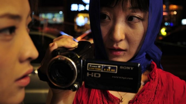 A woman wearing a blue headscarf and a red jacket pointing a video camera at another woman.