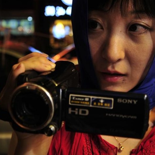 A woman wearing a blue headscarf and a red jacket pointing a video camera at another woman.
