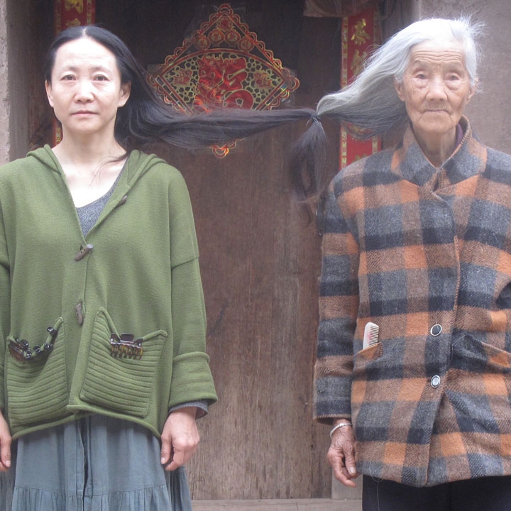 Two women side by side, younger woman with black hair on the left and older woman with white hair on the right. The ends of their long hair are knotted together between them.