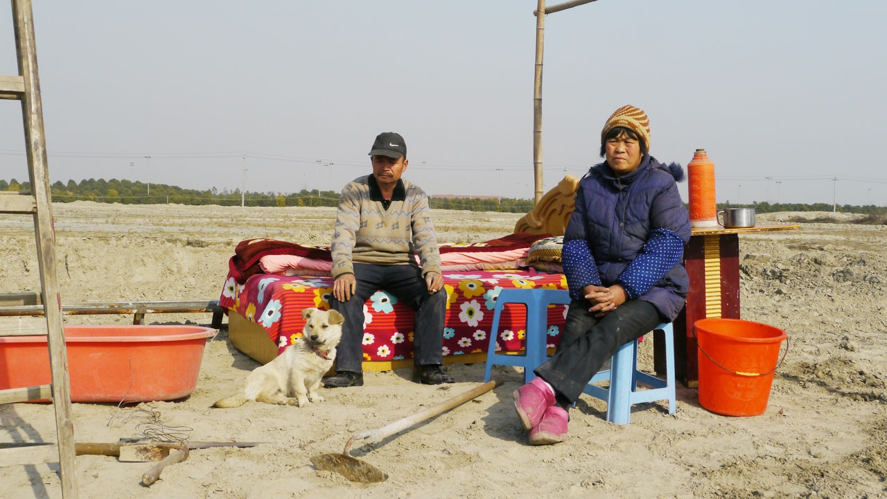 A man sitting on a bed in an open plains. A dog sits at his feet. A woman is sitting on a chair to his left.