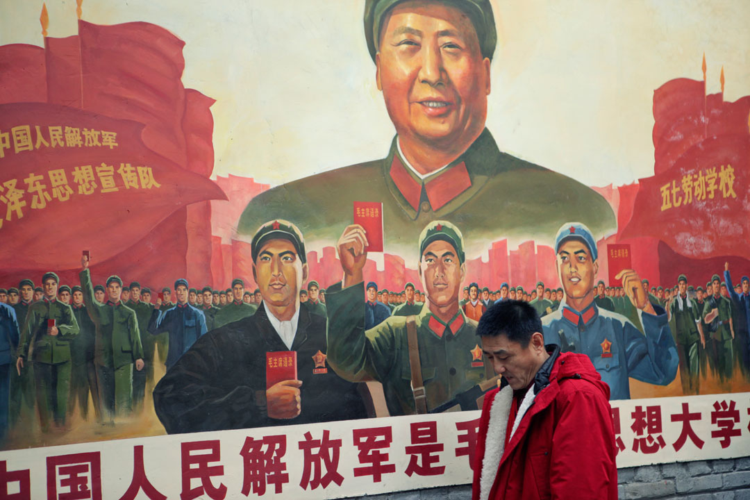 Man in a red sweatshirt walking in front of a poster of Mao Zedong