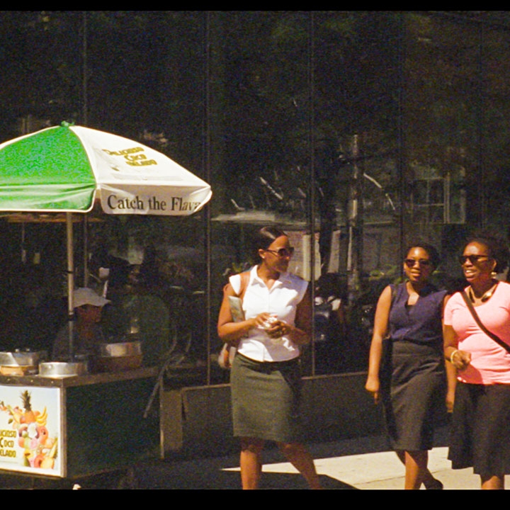 Three women wearing sunglasses, walking down the street on a sunny day, passing an ice cream vendor on the sidewalk.