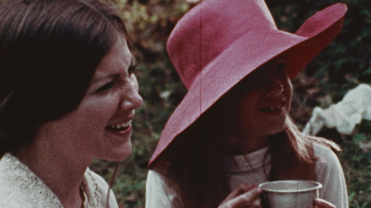 Two women laughing, one with brown hair, another wearing a red hat and drinking tea