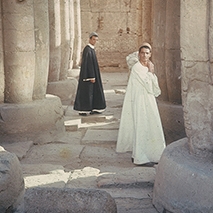 Two figures standing between stone pillars, one in the background wearing black, the one in the foreground wearing white