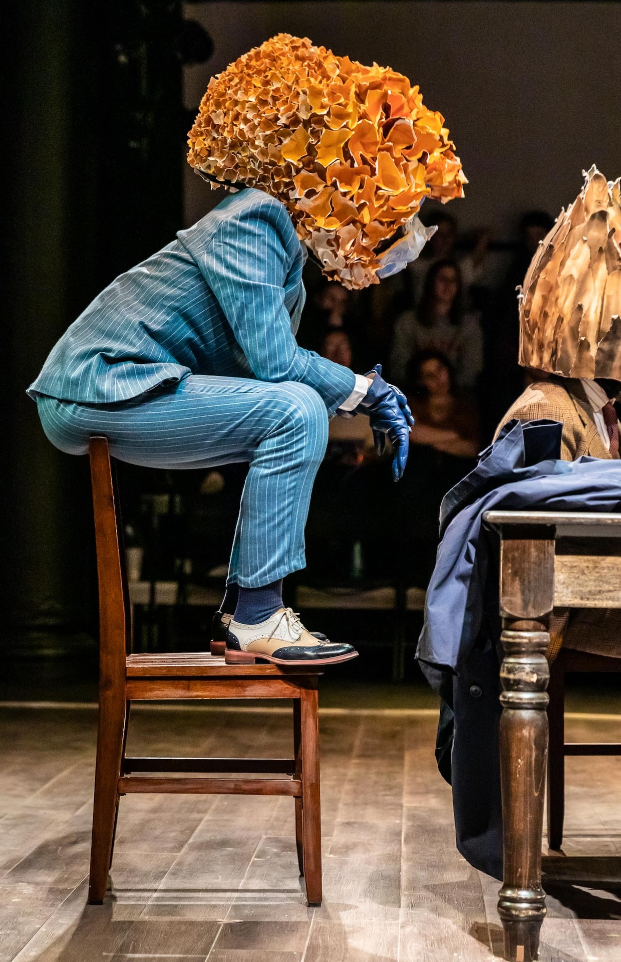 Actor in Blue suit and orange mask sitting on the back of wooden chair
