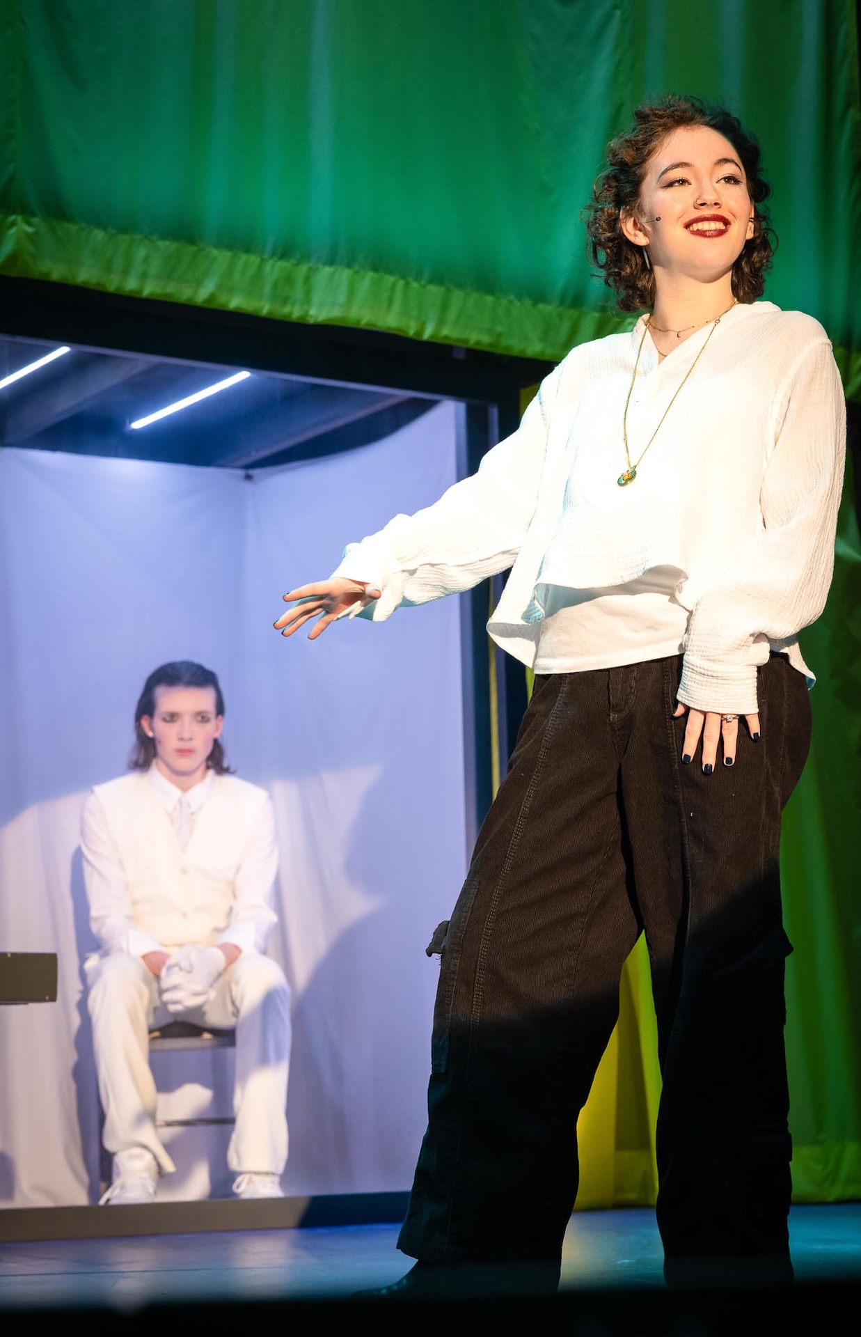 actor standing in foreground wearing white shirt and other actor sitting in background in all white costume