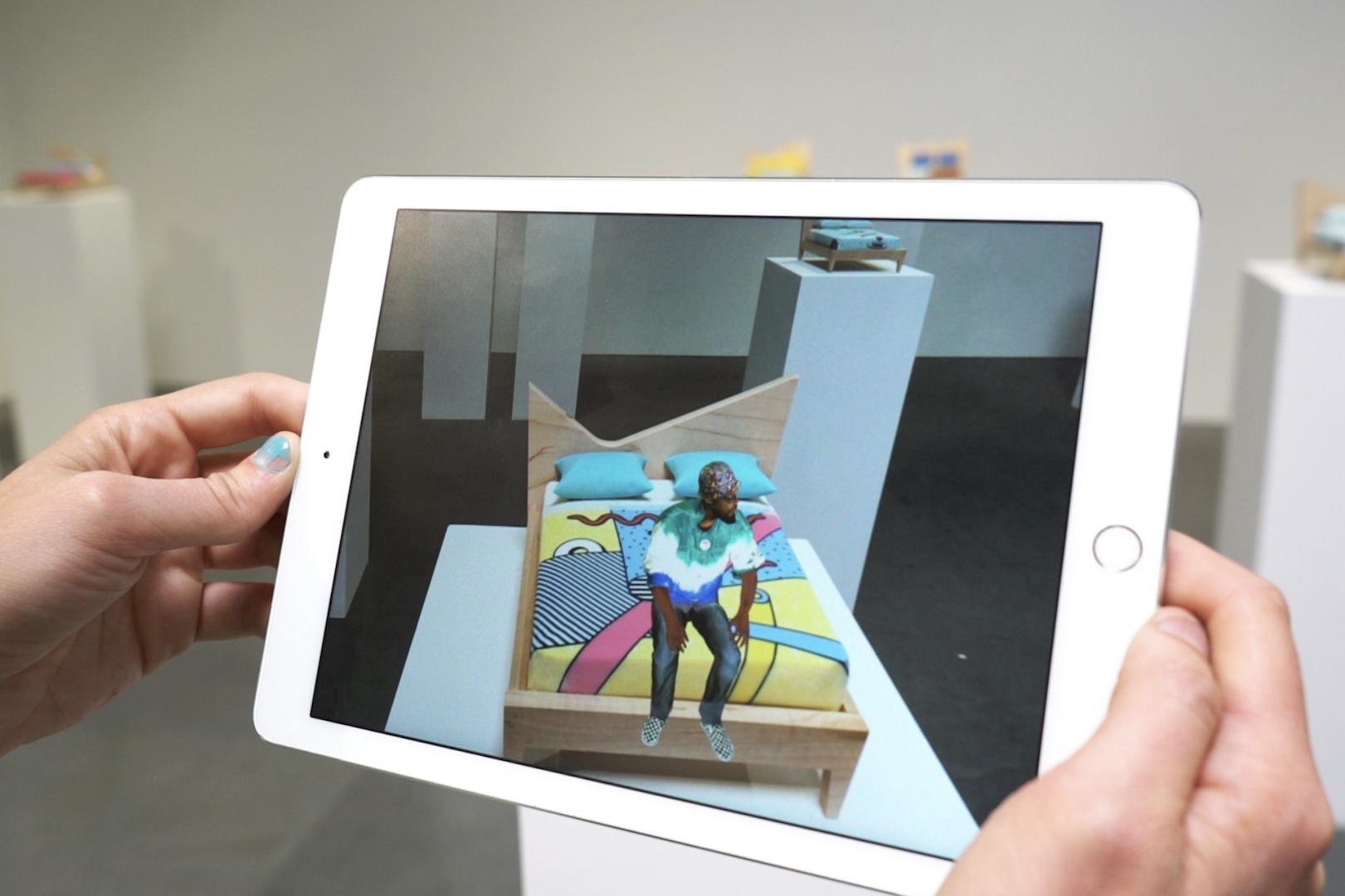Someone holding up an iPad to a museum object of an empty bed, the Augmented Reality shows a person sitting in bed on the screen