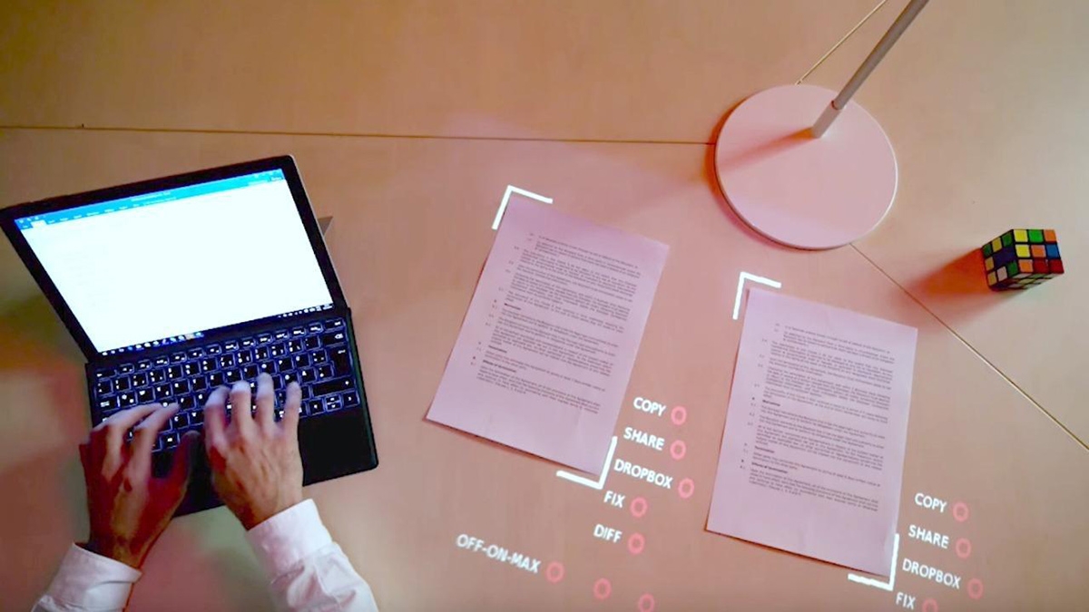 Aerial view of a person typing on a laptop with papers, a lamp and rubic's cube with digital words projected around the papers