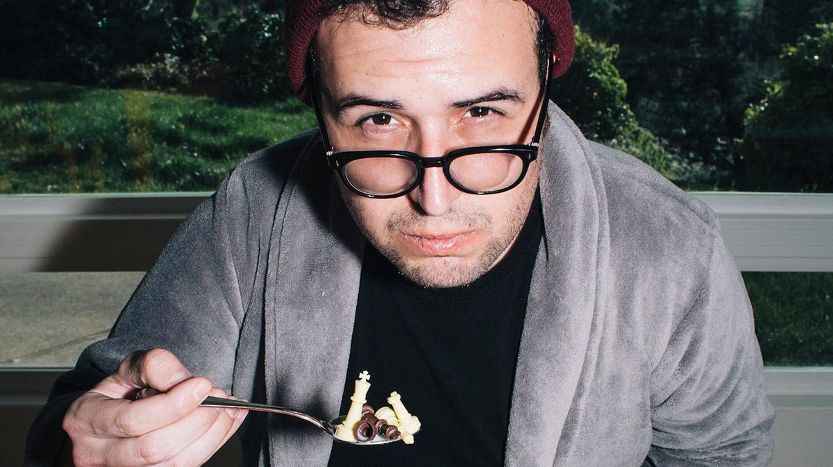 A man looking skeptically over his glasses at the camera, while eating chess pieces in a spoon