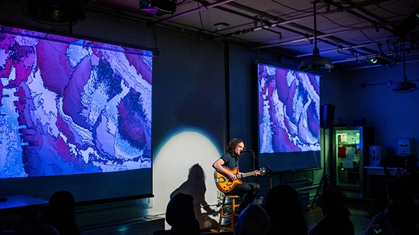 Student plays guitar in the ITP lounge in front of large projections