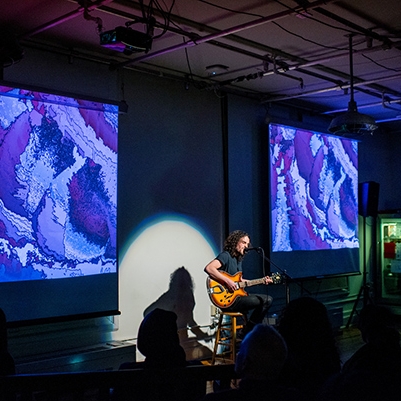 Student plays guitar in the ITP lounge in front of large projections