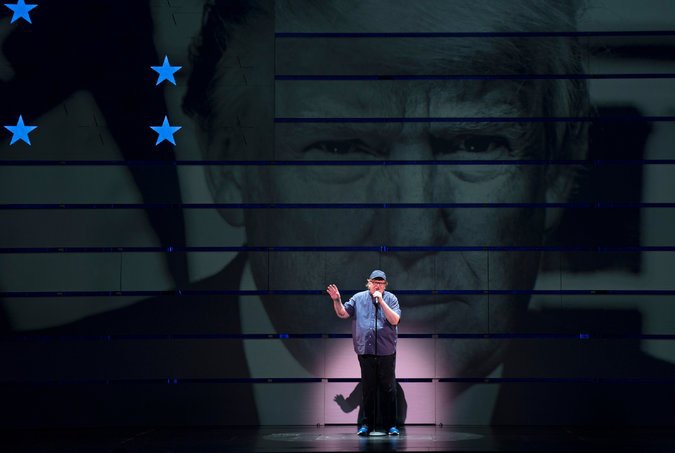 Michael Moore speaking on a big stage against the backdrop of Donald Trump's close up image