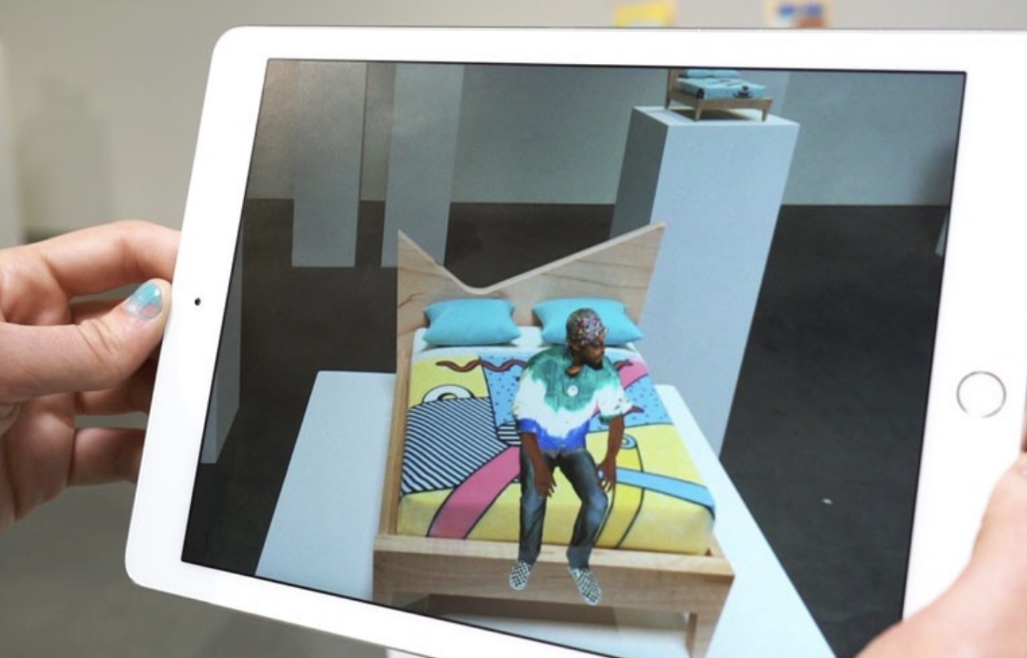 Image of someone holding up an iPad o one of Gabe's installation pieces. One the iPad you see an animated figure sitting on the bed, which isn't present in "real life".