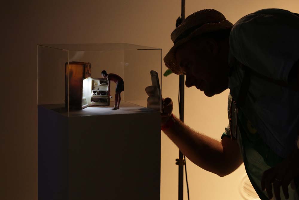 A miniature sculpture of a person looking into a fridge, while a person takes a photo of it