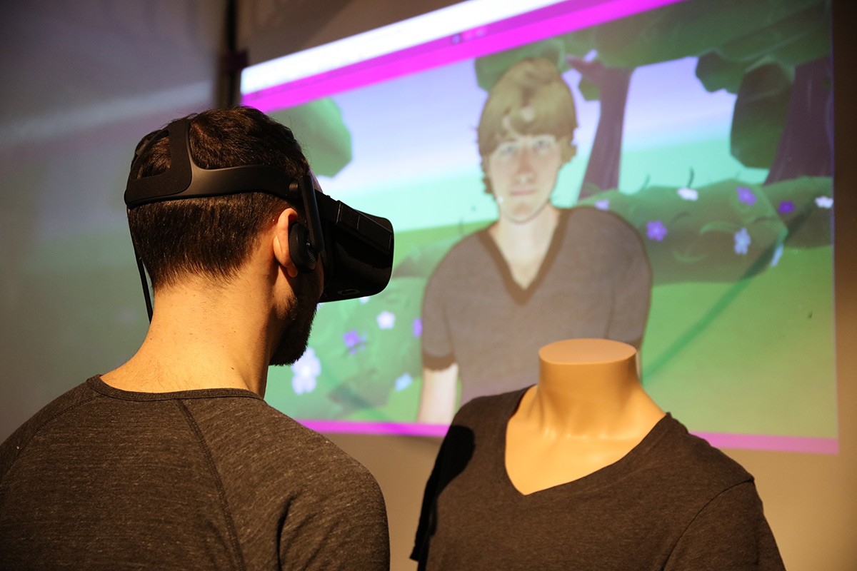 A man wearing VR googles standing next to a headless mannequin with a computer generated image of a person projected