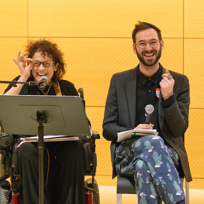 On the left, a white woman in a wheelchair with curly hair adjusts her glasses, smiling. On the right, a bearded white man laughs, mid-snap.