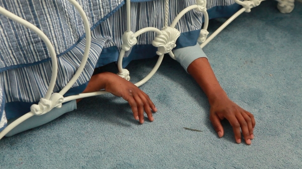 A pair of hands protrudes from beneath a bed, its frame, and comforter. An autobiographical image by Elliot Brown, Jr.