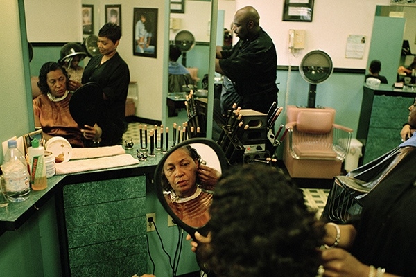 Carrie Mae Weems looks at her reflection in a handheld mirror while sitting for a haircut.
