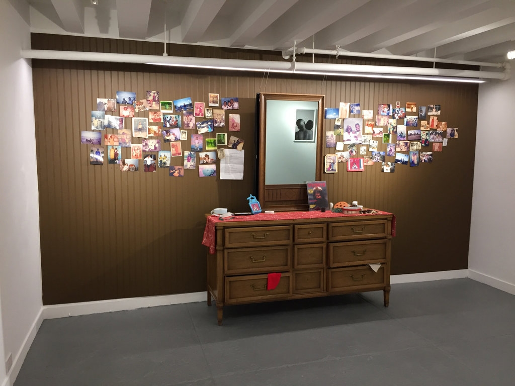 Installation view of Tiger Balm. A bureau and mirror with various photographs stuck to a wood paneled wall.