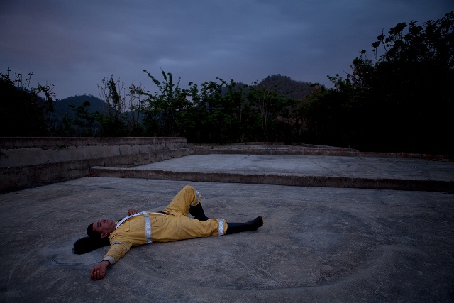 Man lying on the ground with arms outstretched in an open concrete plaza near a field.
