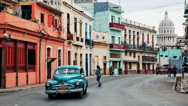 Location shot of a street in Havana showing colorful buildings, a car and people walking.