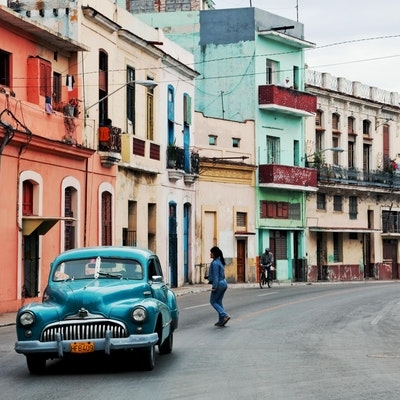 Location shot of a street in Havana showing colorful buildings, a car and people walking.