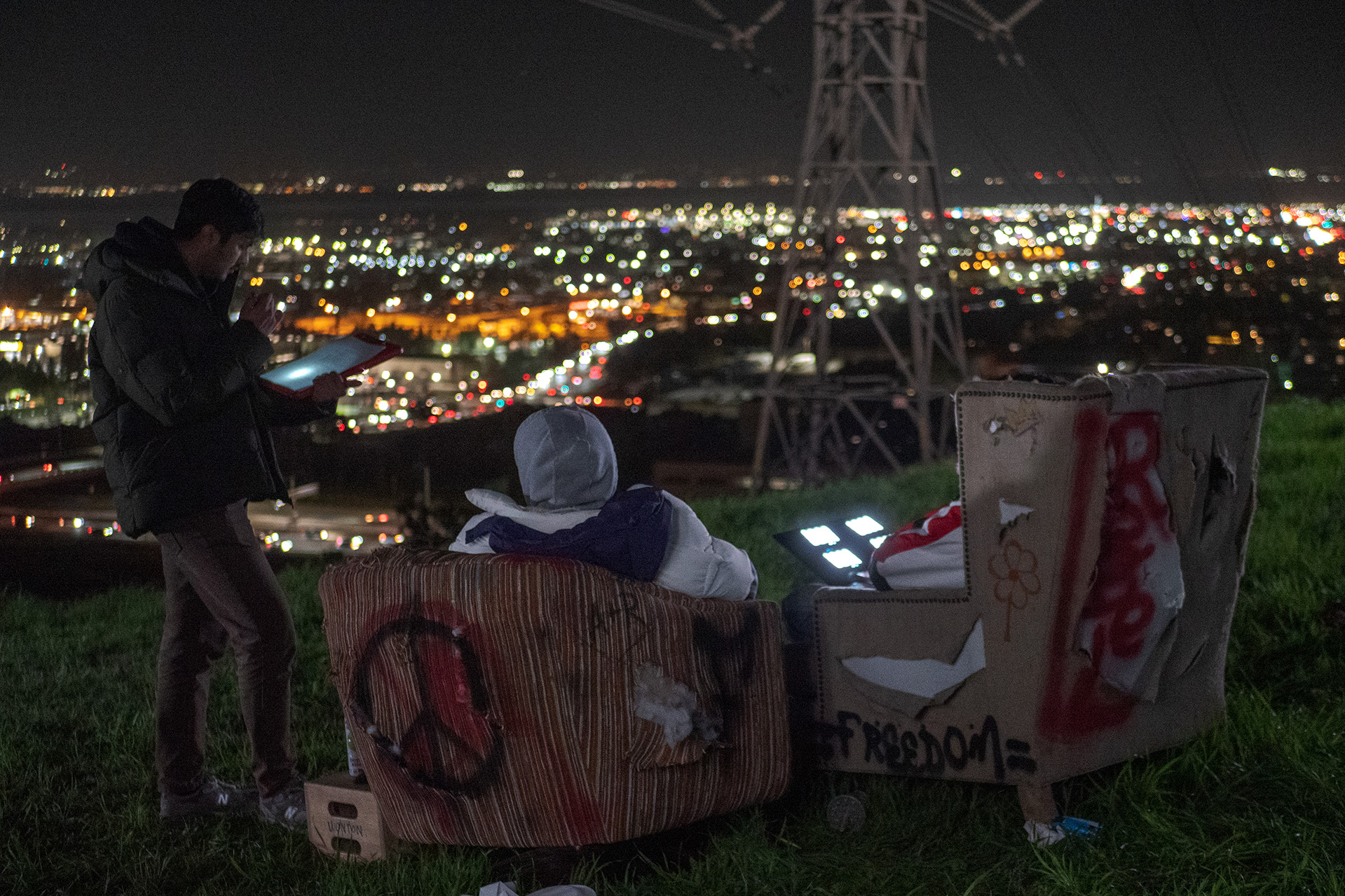 Three young men on ipads, two graffitied chairs, above a city scape at night