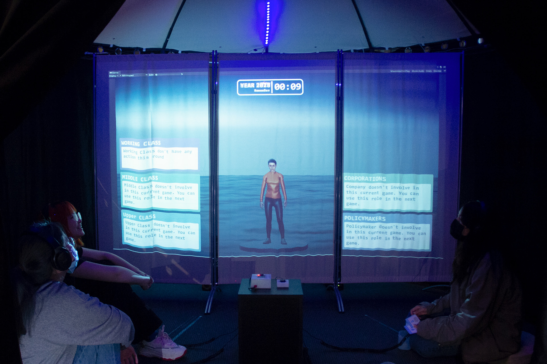 Four people sitting in a tent looking at a projected visual of a 3D avatar
