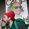Mycah Hogan (above) in a community workshop, RI. Mycah, a man wearing a red hat, red clown nose, green shirt and green turtle shell backpack has his clown nose booped by a small child’s hand. A poster in the background of a colorful Shakespearean era man with glasses, possibly meant to be Shakespeare.