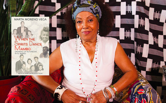 Dr. Marta Moreno Vega sits in a white shirt, bracelets and blue headscarf with hands together. Her book "When the Spirits Dance Mambo" cover is placed over her arm in the image