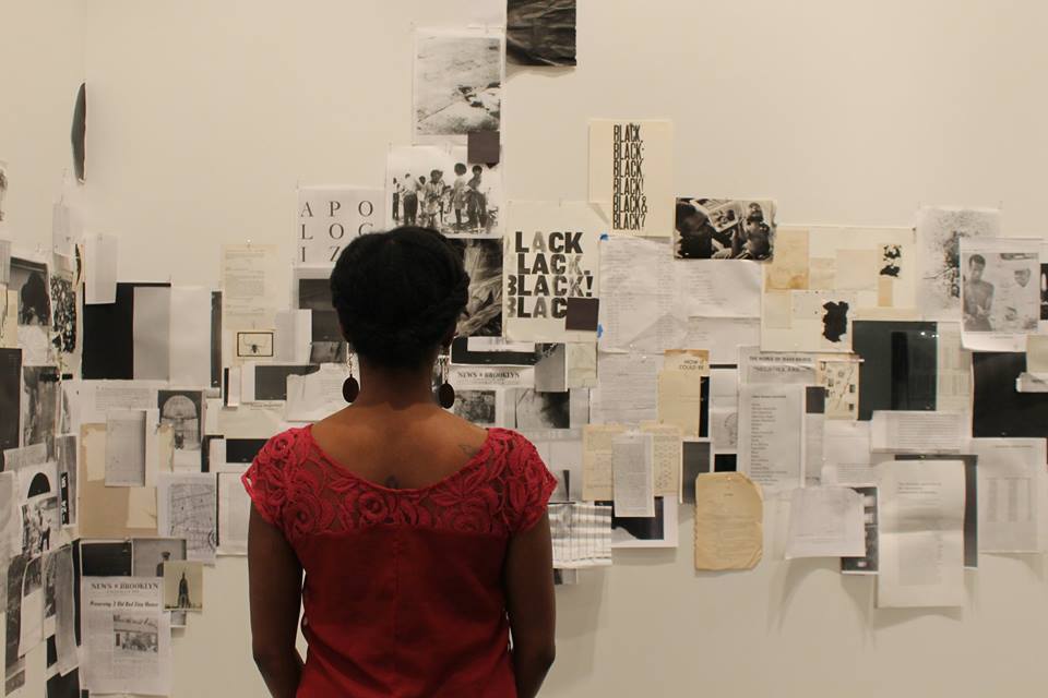 Black woman wearing red shirt, back to audience, looks at papers taped to a white wall