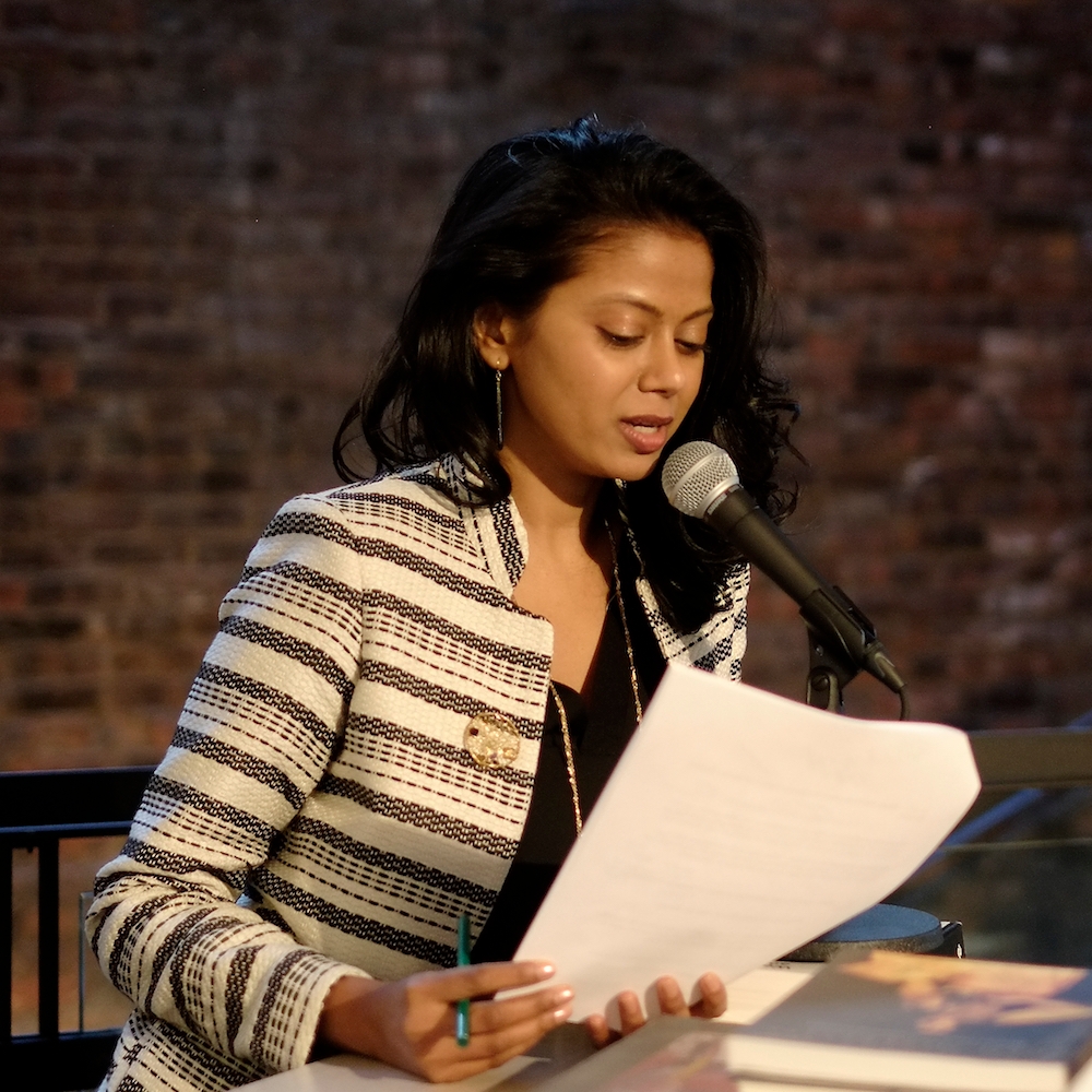 Grace Ali wears brown striped jacket, holding papers and speaking at a microphone