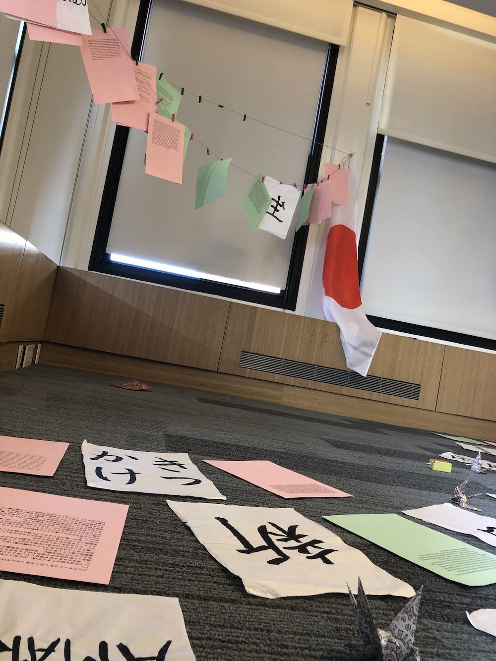 Installation in the corner of a room with a clothesline with pink and green papers with text and Japanese characters hang and are also scattered on the floor