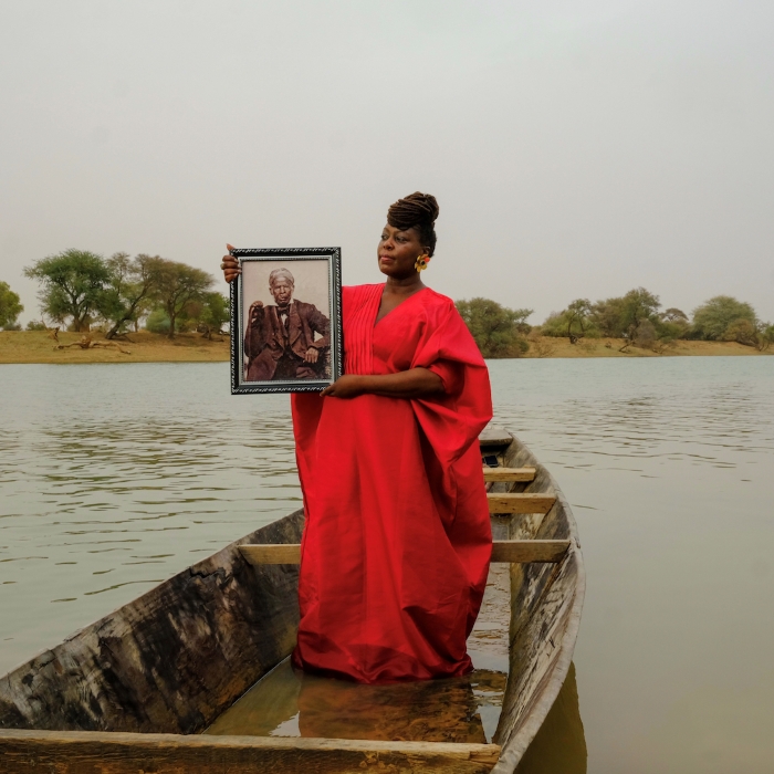 Black woman wears blue long sleeve top and black hat and glasses and poses facing left with hand on hip and head tilted toward camera