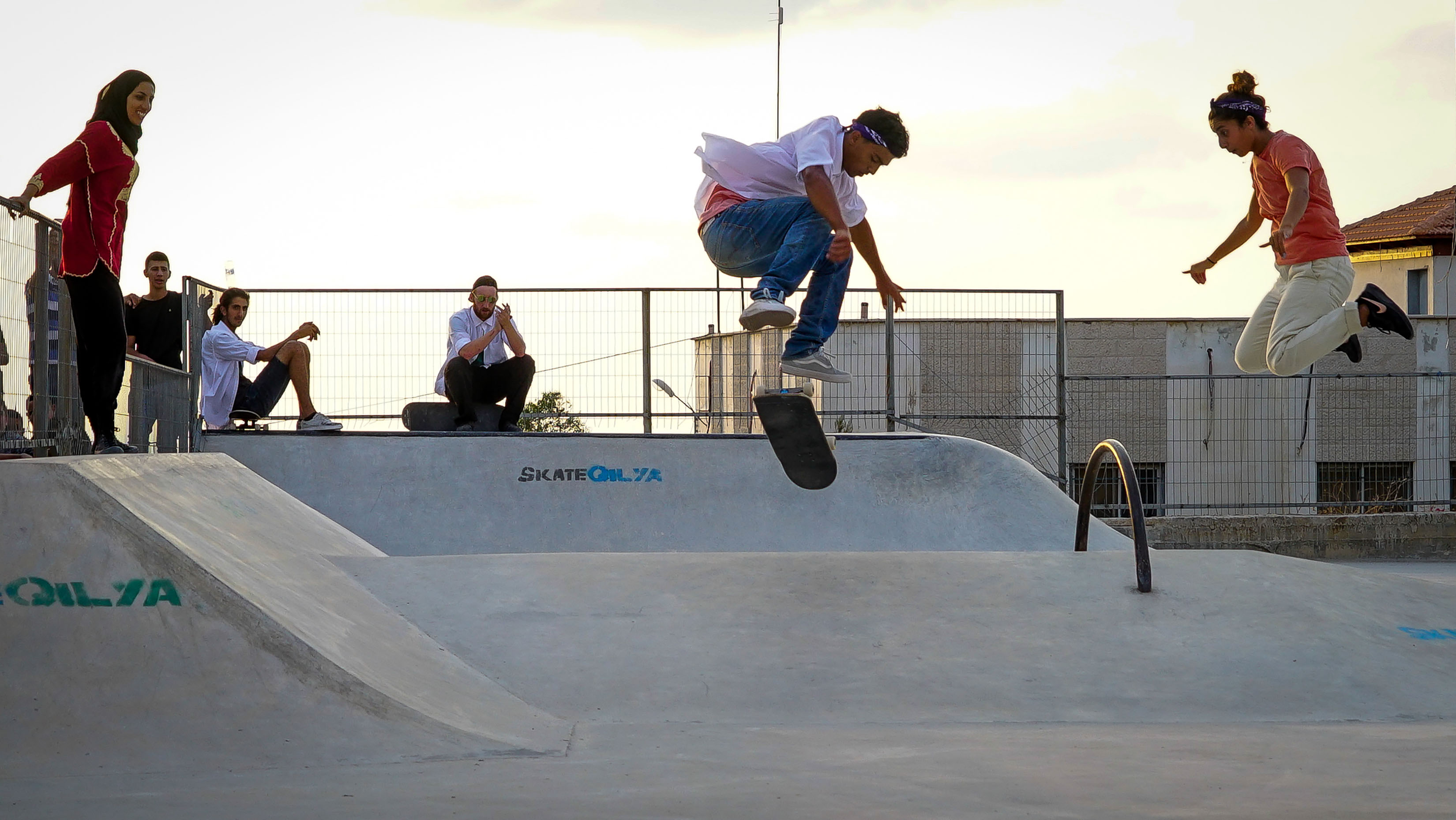 A photo from "A Skate Play." There is a woman with a hijab standing on the left, a man skateboarding in the center, and a woman jumping on the right.