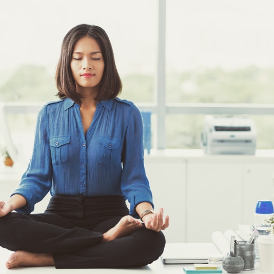 A woman sitting on an office desk meditating in lotus position.