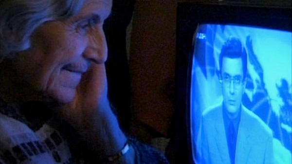 An elderly woman leaning in closely to look at a television screen, a man in a suit on the screen.