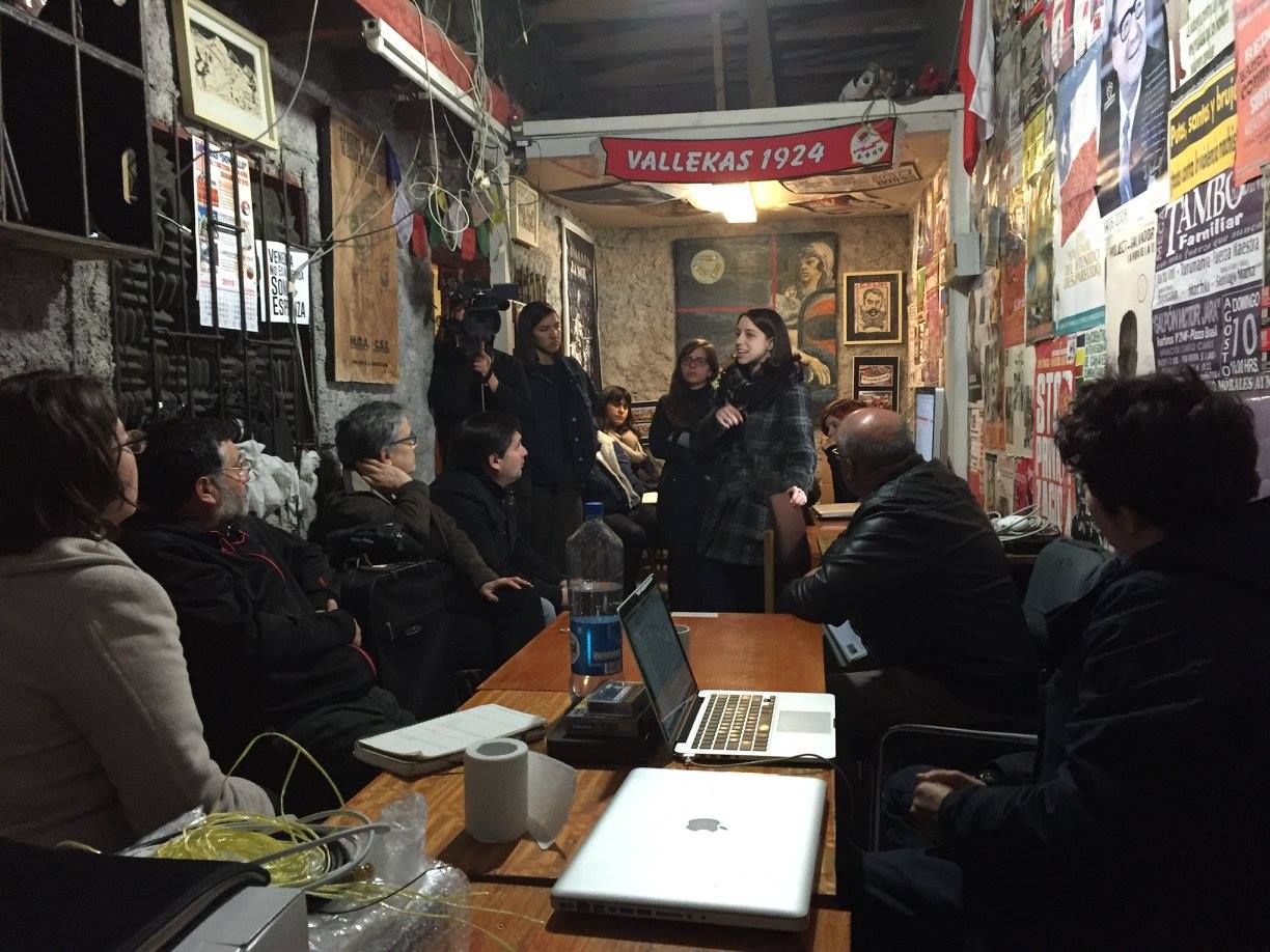 A woman addressing a room of people, posters on the walls and a flag behind her.