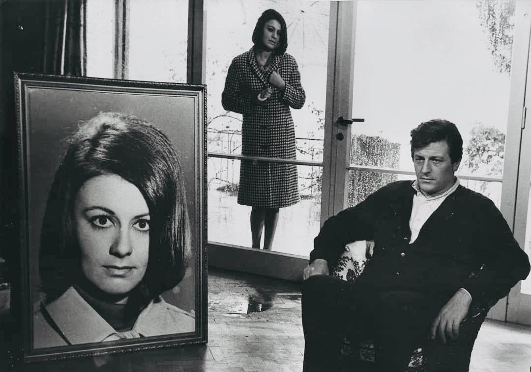 A man sitting in a chair looking at an enlarged, framed photo of a woman. A woman stands behind both.