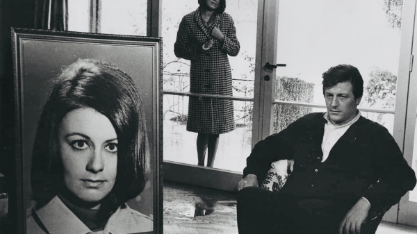A man sitting in a chair looking at an enlarged, framed photo of a woman. A woman stands behind both.