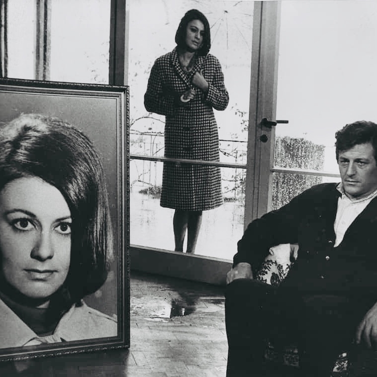 A man sitting in a chair looking at an enlarged, framed photo of a woman. A woman stands behind both.