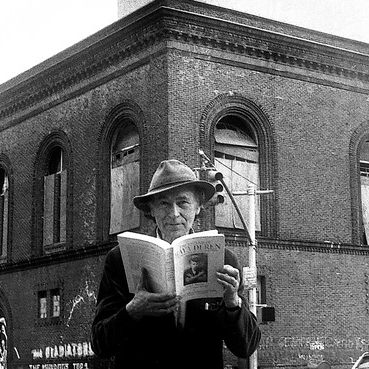 Filmmaker Jonas Mekas, wearing a hat and reading a book, standing outside of Anthology Film Archives building.