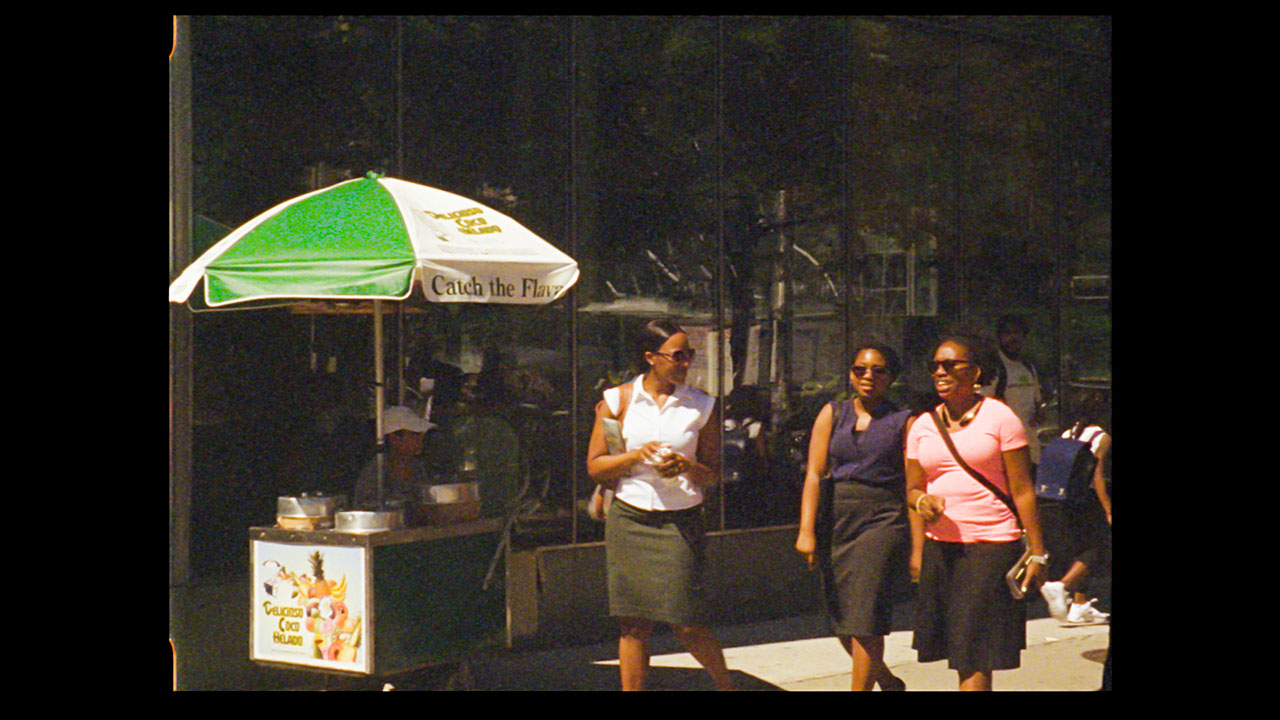 Three women wearing sunglasses, walking down the street on a sunny day, passing an ice cream vendor on the sidewalk.
