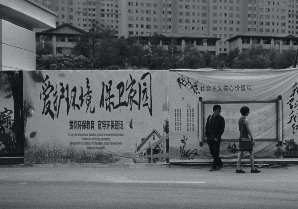 Man and woman passing each other on the street, with propaganda in chinese characters in the background