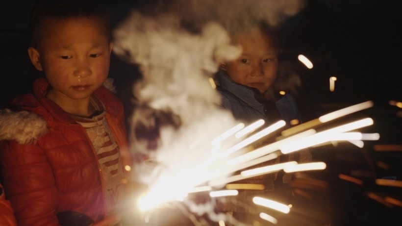 Two children, one in the foreground wearing a bubble coat with a sparkler in the the foreground