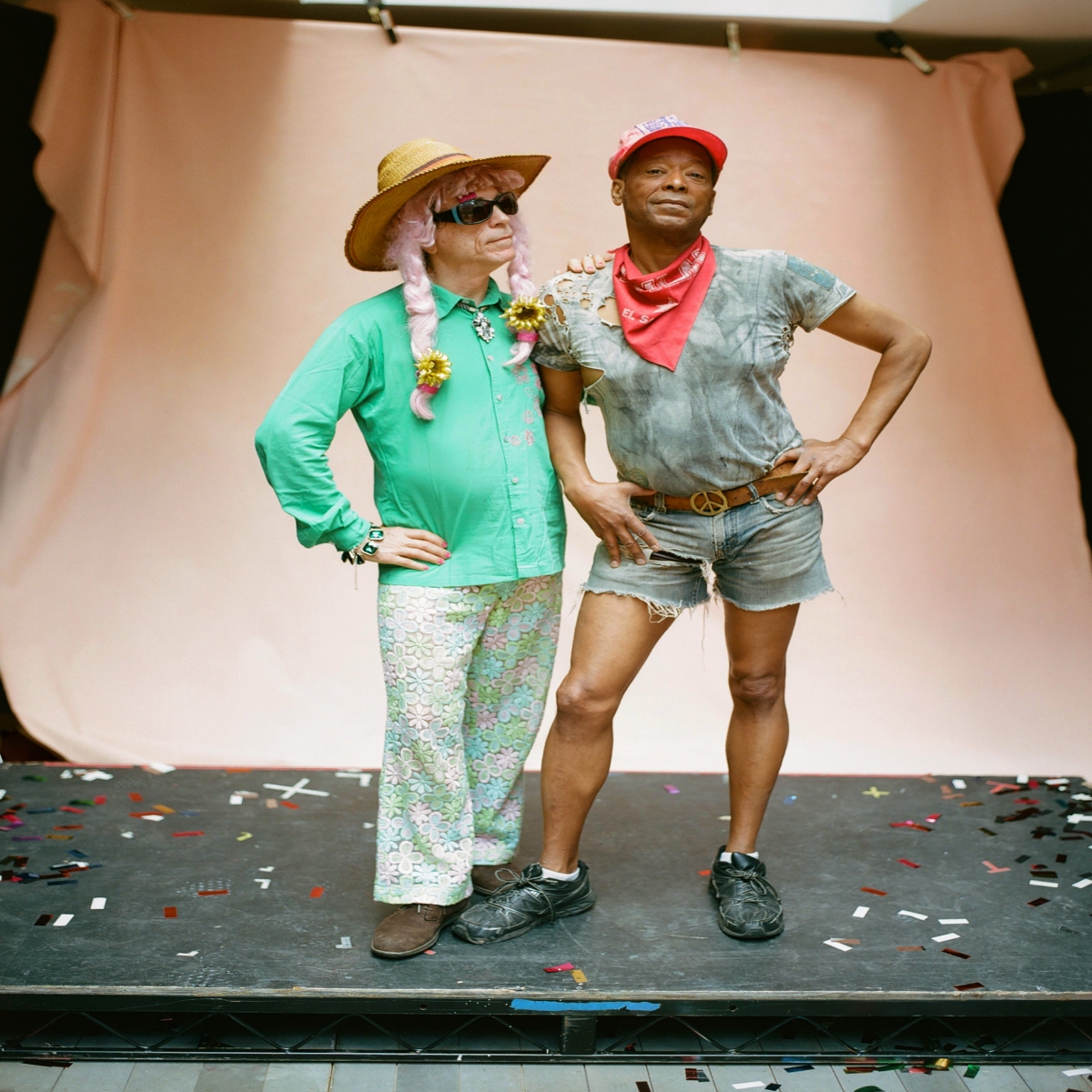 Two men standing next to each other. One, wearing a cowboy hat and a teal shirt with floral pants and the other wearing a red cap, red kerchief, and denim shorts. 